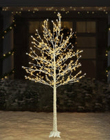 This is a lifestyle image showing the birch tree decoration on the snowy ground in front of a house. The lights are warm white and pure white.