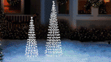 This animated image shows two white wire cone trees twinkling on a snowy lawn in front of a house decorated for Christmas.