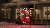 This animated image shows this twinkling candy cane decoration on a snowy yard in front of a house decorated for Christmas.