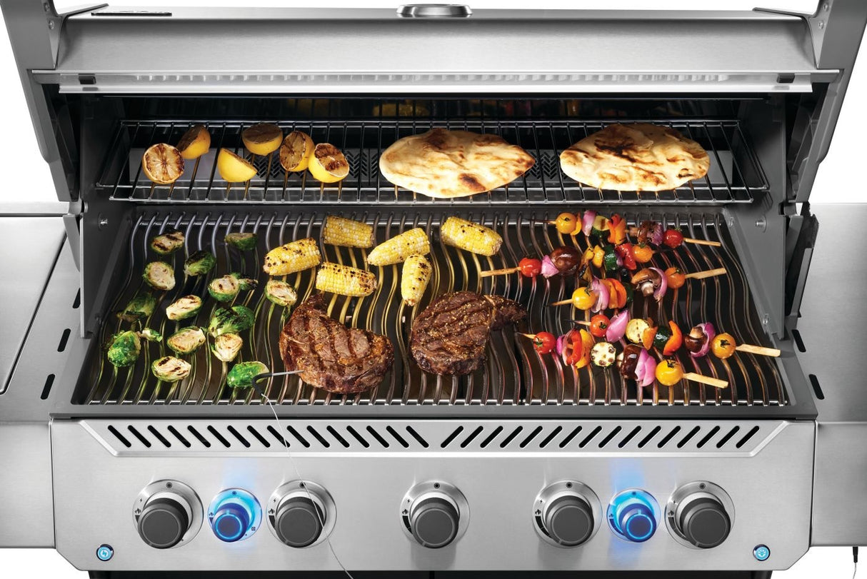 This is an up close view of food cooking on the grill. There are lemons and tortillas cooking on the top shelf. There are Brussel sprouts, corn on the cob, kebabs and two steaks cooking on the bottom shelf. 