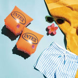 This is a lifestyle image showing these two arm floaties on a blue background with a pair of small sunglasses, a small hair clip, a yellow towel and a pair of small white and blue striped shorts.