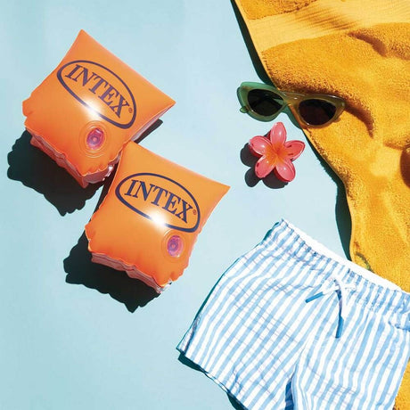 This is a lifestyle image showing these two arm floaties on a blue background with a pair of small sunglasses, a small hair clip, a yellow towel and a pair of small white and blue striped shorts.