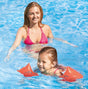Neon orange colored inflatable arm bands for children. The inflatable goes around the two upper arm areas to help them stay afloat in water. This image shows a small child, happily swimming with her mom nearby.