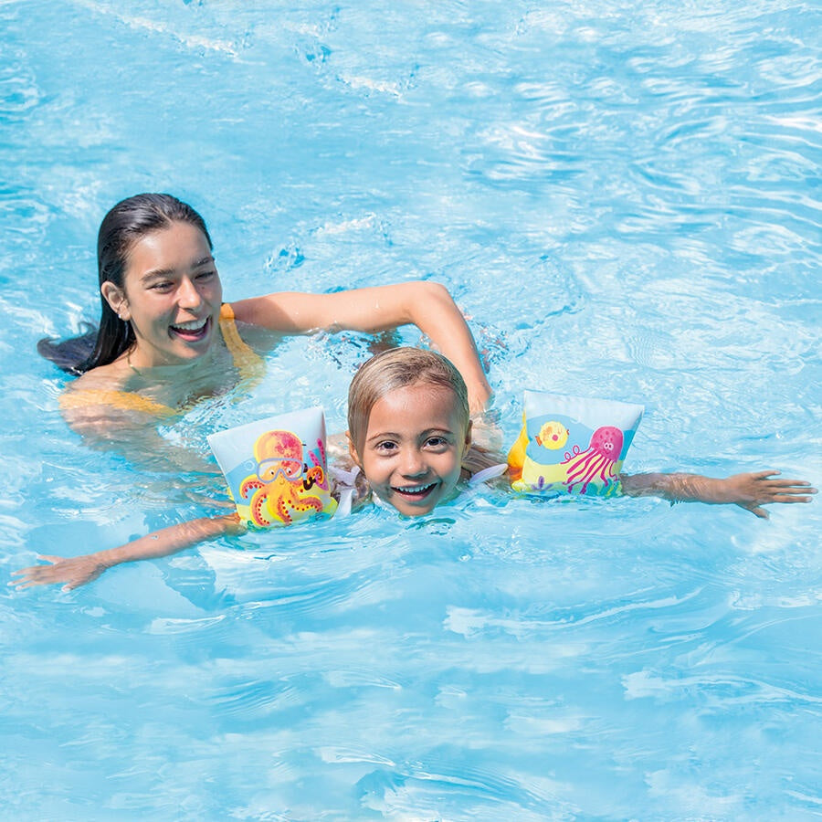 This is a lifestyle image showing a young girl swimming with the arm bands on it while her mom helps from behind.