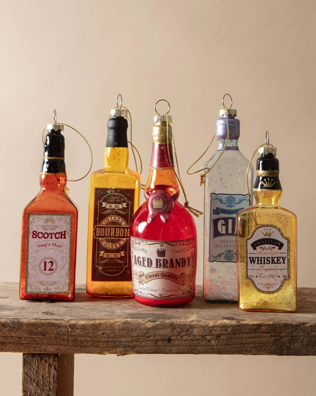 Ornament shaped like a miniature bottle of alcohol. One ornament is red and reads "Aged Brandy." One ornament is yellow and reads "Whiskey." One ornament is orange and reads "Scotch." One ornament is yellow and reads "Bourbon." One ornament is light blue and reads "Gin." Each ornament has glitter inside.