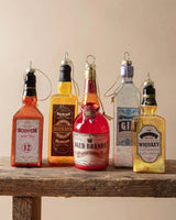 Ornament shaped like a miniature bottle of alcohol. One ornament is red and reads "Aged Brandy." One ornament is yellow and reads "Whiskey." One ornament is orange and reads "Scotch." One ornament is yellow and reads "Bourbon." One ornament is light blue and reads "Gin." Each ornament has glitter inside.