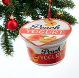 Ornament shaped like a cup of yogurt. One ornament is white and red and "flavored" strawberry. One ornament is white and blue and "flavored" blueberry. One ornament is white and orange and "flavored" peach. One ornament is white and blue and "flavored" Greek-style.