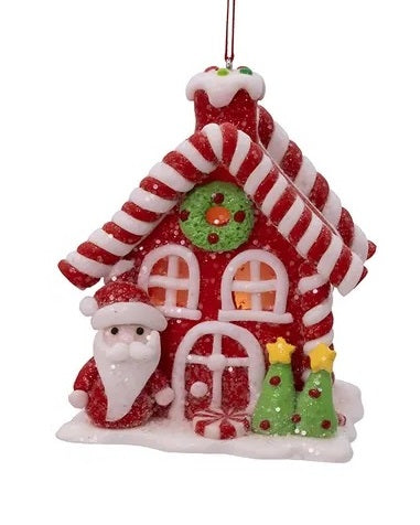 Ornament shaped like a house made out of candy. This one is a red house adorned with candy canes, gumdrops, lollipops, frosting and other sweet treats. Outside of the house there are two Christmas trees and Santa Claus.