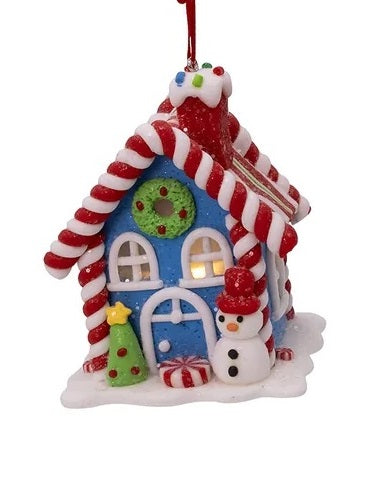 Ornament shaped like a house made out of candy. This one is a blue house adorned with candy canes, gumdrops, lollipops, frosting and other sweet treats. Outside of the house there's a Christmas tree and a snowman wearing a red hat.