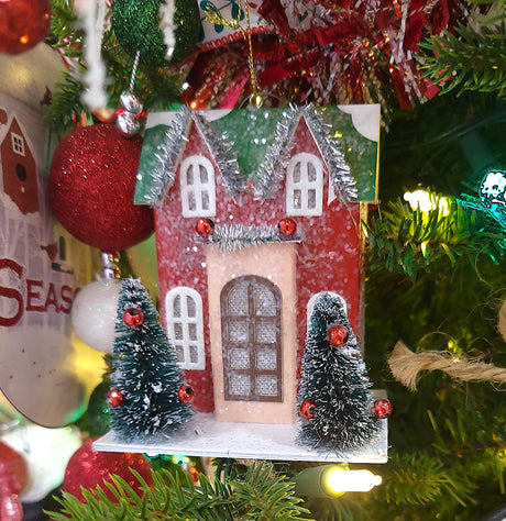 Christmas ornament shaped like a house. Colors are red, green, tan and white.