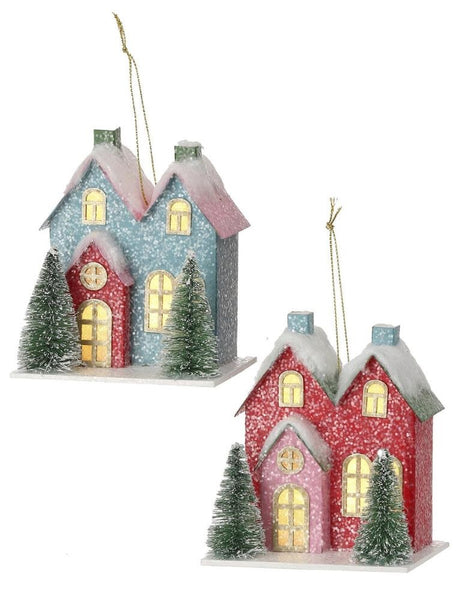 Christmas ornament shaped like a cute house with two trees in the front. One is blue with a red door and one is red with a pink door. LED lights are visible inside from the windows.