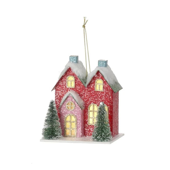 Christmas ornament shaped like a cute house with two trees in the front. One is blue with a red door and one is red with a pink door. LED lights are visible inside from the windows.