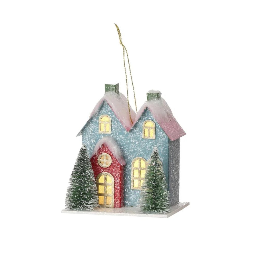 Christmas ornament shaped like a cute house with two trees in the front. One is blue with a red door and one is red with a pink door. LED lights are visible inside from the windows.