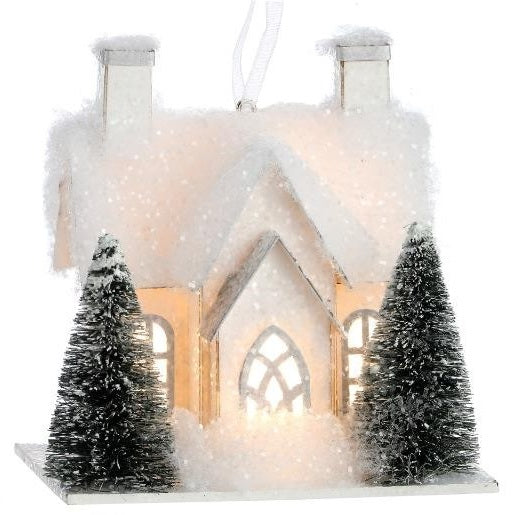 Ornament designed to look like a small house with three windows and two chimneys. Both homes are covered in white glitter and fluff and have two snow-covered trees in the front yard.