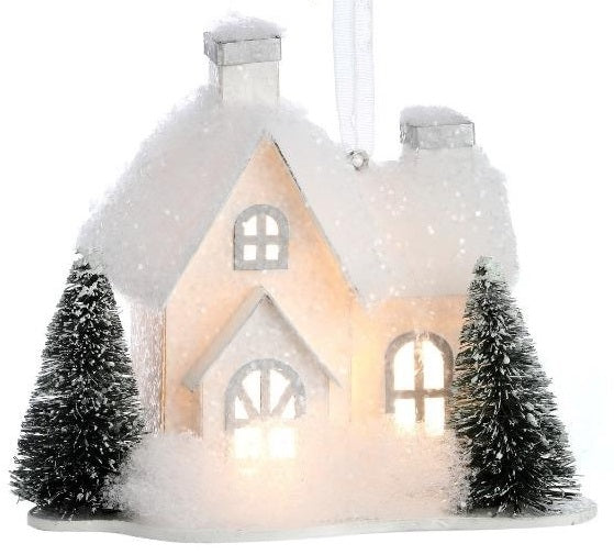 Ornament designed to look like a small house with three windows and two chimneys. Both homes are covered in white glitter and fluff and have two snow-covered trees in the front yard.