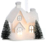 Ornament designed to look like a small house with three windows and two chimneys. Both homes are covered in white glitter and fluff and have two snow-covered trees in the front yard.