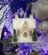 Ornament designed to look like a small house with three windows and two chimneys. Both homes are covered in white glitter and fluff and have two snow-covered trees in the front yard. There are two different options.