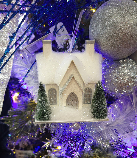 Ornament designed to look like a small house with three windows and two chimneys. Both homes are covered in white glitter and fluff and have two snow-covered trees in the front yard. There are two different options.