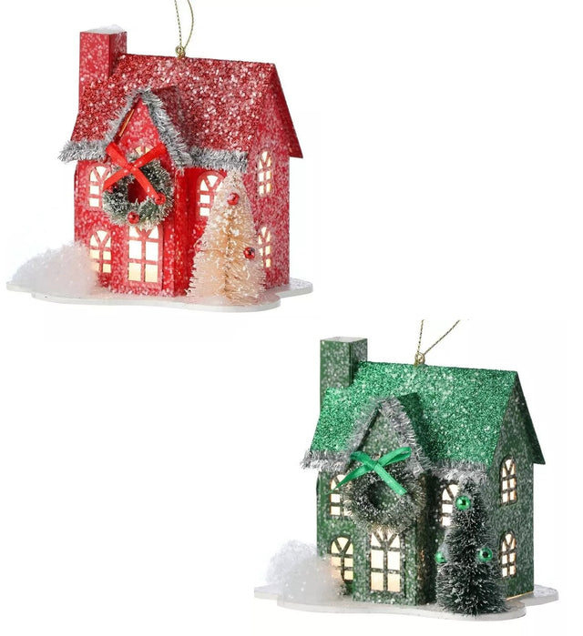 Ornament shaped like a house covered in snow. One house is red and features a wreath near the roof with a red bow. One house is green and features a wreath near the roof with a green bow.