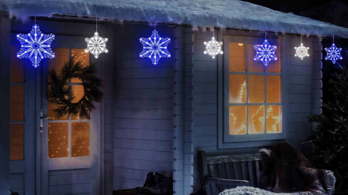 A string of blue and pure white twinkling snowflake icicle lights hanging off the snowy roof of a house.