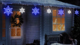 A string of blue and pure white twinkling snowflake icicle lights hanging off the snowy roof of a house.