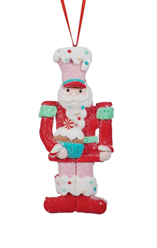 This ornament features a toy soldier wearing a red jacket, pink pants, a pink hat and is holding a blue cupcake.
