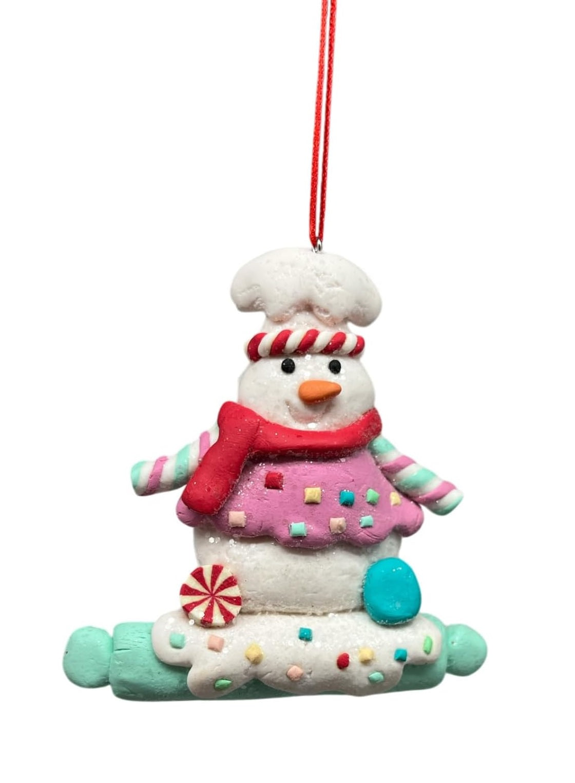 This image shows a snowman wearing a chef's hat. He's wearing a red scarf and a pink sweater and he's standing on a light blue rolling pin.