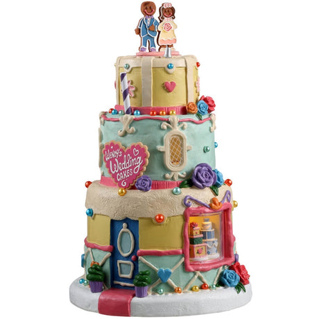 Resin lighted building for a Lemax Village display. Designed to look like a three tiered wedding cake. There's a gingerbread cookie couple on top and a cookie-shaped sign that reads "Wendy's Wedding Cakes." The top tier is yellow, the middle tier is a light teal color and the bottom tier is both yellow and light teal. There are sprinkles and frosting accents all over.