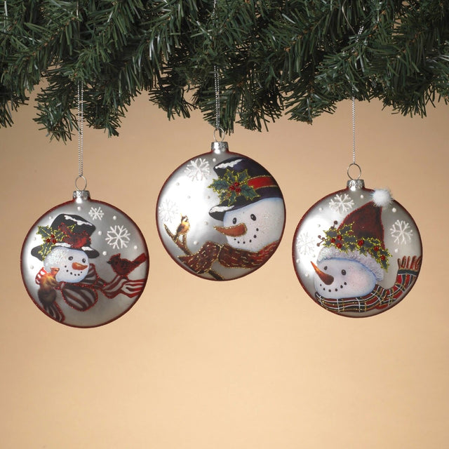 Round ornament featuring a snowman in a winter scene. One ornament has a snowman wearing a red and white scarf and a top hat and is smiling at a squirrel and two cardinals. One ornament has a snowman wearing a red and gold scarf and a top hat and is smiling at a bird on a branch. One ornament has a snowman wearing a red, green and gold scarf and a Santa hat. Each ornament features holly leaves and berries and snowflakes.