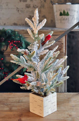 Tabletop tree in a birch planter box. The branches of the tree are dusted with white to mimic snow. The tree is accented with two red cardinal birds and some snow-covered pine cones.