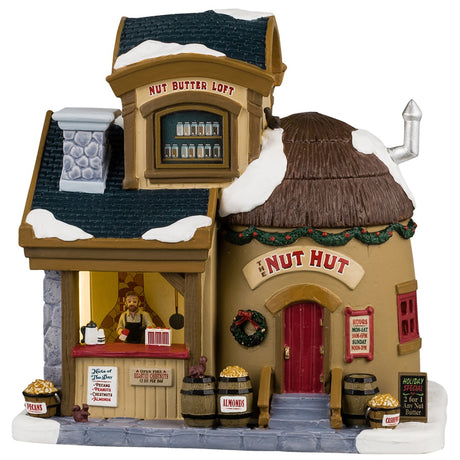 Porcelain lighted building for a Lemax Village display. Designed to look like a store booth connected to a building that is shaped like a coconut. There's snow on the roof and the sidewalk. There's a man behind the booth wearing an apron. On the upper level of the building, there's a Nut Butter Loft. There's a decorated wreath and garland near the front door. There are barrels of different nuts and a few squirrels.