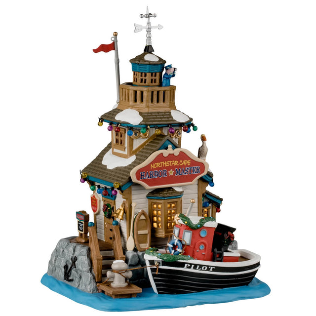 Porcelain lighted building for a Lemax display. Designed to look like a building next to a body of water. There's someone using binoculars on the balcony on the roof. There are multi-colored lights and a pelican sitting near the sign. The sign reads "Northstar Cape Harbor Master." There's a boat tied up at the dock. The boat has the word "PILOT" written on the side and has a decorated garland around it.