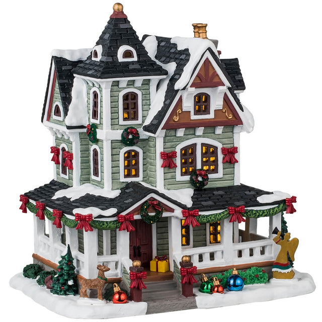 Porcelain lighted building for a Lemax display. Designed to look like a Victorian-era house covered in snow. It's decorated with decorated wreaths, garland, large shiny ornaments on the ground, Christmas trees and bushes and an angel silhouette.