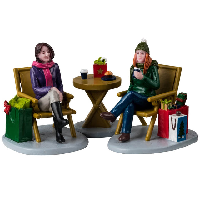 Resin figurines for a Lemax Village tabletop display. Designed to look like two women enjoying a cup of coffee. They're both sitting in chairs, there's a small, round table in between them. They're both wearing winter clothes and have shopping bags next to them.