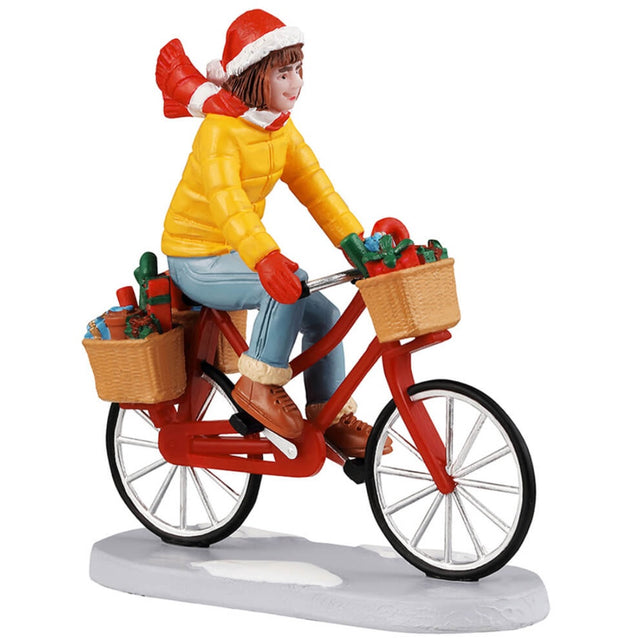 Resin figurine for a Lemax display. Designed to look like a girl wearing a Santa hat, a red and white scarf, a yellow jacket, red mittens, jeans and UGG like boots. She's riding a red bike with three baskets full of presents. There's some snow on the ground.