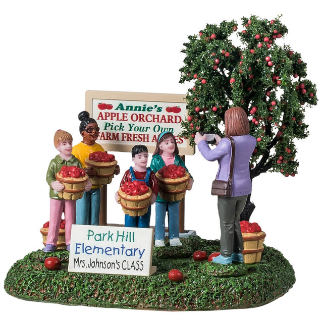 Resin table accent for a Lemax Village tabletop display. Designed to look like four students posing for a picture. They're standing next to an apple tree and a sign that reads "Annie's Apple Orchard. Pick your own farm fresh apples." The children are all smiling and holding a basket of apples. There are apples on the ground and a sign that reads "Park Hill Elementary - Mrs. Johnson's class." Mrs. Johnson is the one taking a picture of the students. The ground is covered in grass.