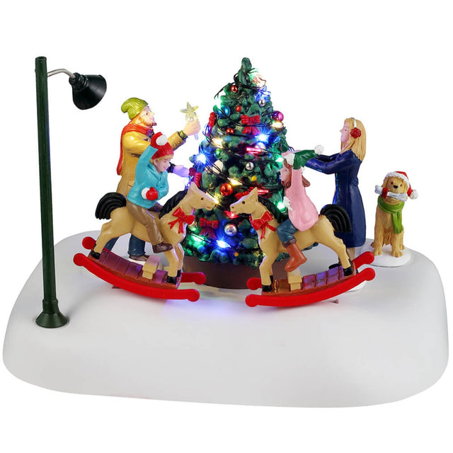 Animated resin village accessory for a Lemax display. Designed to look like a family decorating a Christmas tree on a snowy patch. They're all wearing winter clothes. The kids are riding on reindeer that rock back and forth. There's also a dog watching, wearing a Santa hat. There's a pole nearby with a light on it.