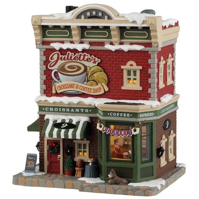 Porcelain lighted building for a Lemax tabletop display. Designed to look like a red, gray and brown brick building. There's snow on the roof and the sidewalk. There are signs advertising croissants, coffee, espresso and sandwiches. Inside the window you can see a man sitting at a table. There's a dog sitting under the window on the sidewalk.