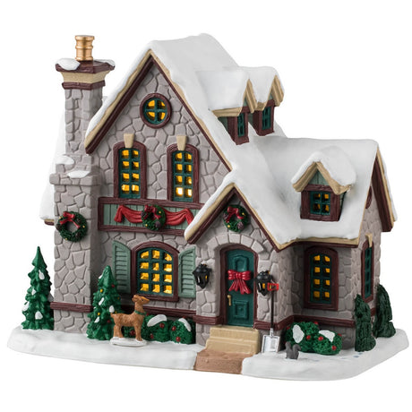 Porcelain lighted building for a Lemax display. Designed to look like a charming, stone cabin covered in snow. There are decorated wreaths and red bows everywhere. There are snow covered trees and bushes around the property. There's a deer looking into the window and a squirrel standing next to a shovel.