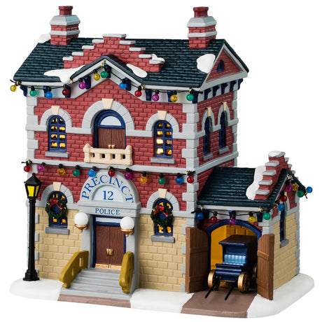 Porcelain lighted building for a Lemax display. Designed to look like a red, yellow and gray brick police precinct office. The roof and sidewalk has snow on it. There are multi-colored lights hanging from the roof. The windows have wreaths and there's a car coming out of the side garage. 