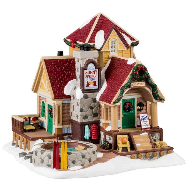 Porcelain lighted building for a Lemax display. Designed to look like a wooden cottage with a stone chimney and a stone hot tub on the porch. There's snow on the building and features wreaths and garland accents. There's a balcony with a chair and a table. There are coats hanging on the side and a few pairs of boots. There's a dog sleeping on the porch and a few pairs of skis leaning up against the stone hot tub.