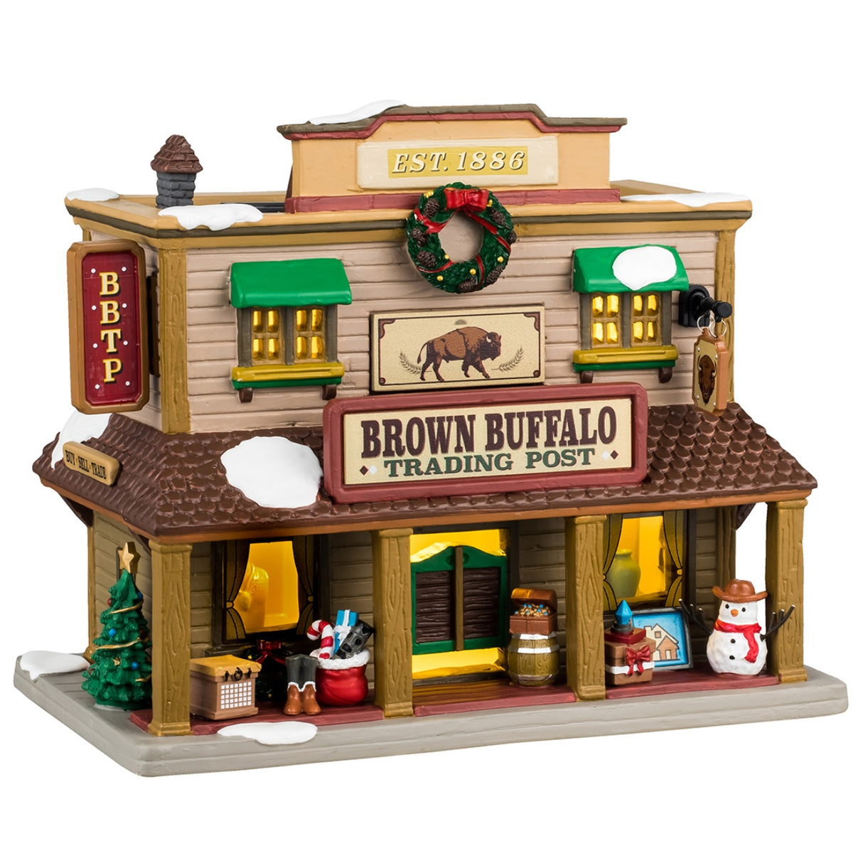 Porcelain lighted building for a Lemax display. Designed to look like a Western building. On top of the building, it says "Est. 1886." There's a large wreath near the roof and a picture of a buffalo. There's snow on the roof and a bunch of Christmas accents throughout. Accents include: a decorated Christmas tree, a Santa bag with presents and a candy cane inside, there are presents and a snowman wearing a cowboy hat.