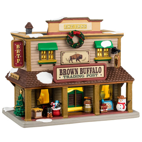 Porcelain lighted building for a Lemax display. Designed to look like a Western building. On top of the building, it says "Est. 1886." There's a large wreath near the roof and a picture of a buffalo. There's snow on the roof and a bunch of Christmas accents throughout. Accents include: a decorated Christmas tree, a Santa bag with presents and a candy cane inside, there are presents and a snowman wearing a cowboy hat.
