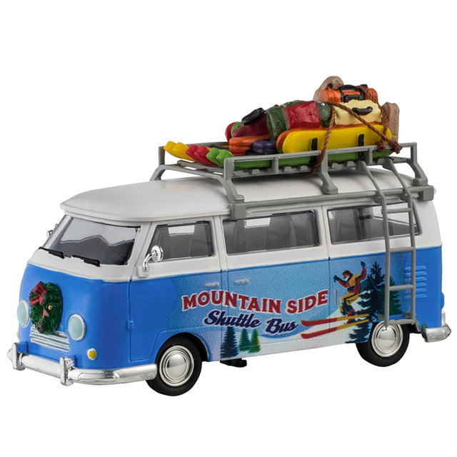 Plastic table accent for a Lemax display. Designed to look like a blue and white Volkswagen shuttle bus. The sides of the bus read "Mountain Side Shuttle Bus" and features a mural of someone skiing down a snowy hill surrounded by trees. There's a wreath on the front bumper. There's a ladder on the side leading up to the roof o the bus that's holding a bunch of ski equipment and luggage.