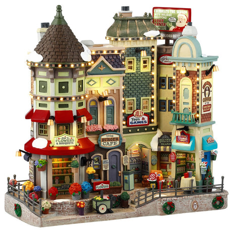 Resin building facade for a Lemax display. Designed to look like a bunch of store fronts. Signs read: "Fay's Flowers & Bouquets," "The Burger Cafe," "Jolly Toys & Games," "Quilter's Island," Sweet Spot" and "Hot Fresh Coffee at the Christmas Bazaar!" There are flowers everywhere and a billboard on the roof advertising a visit with Santa.