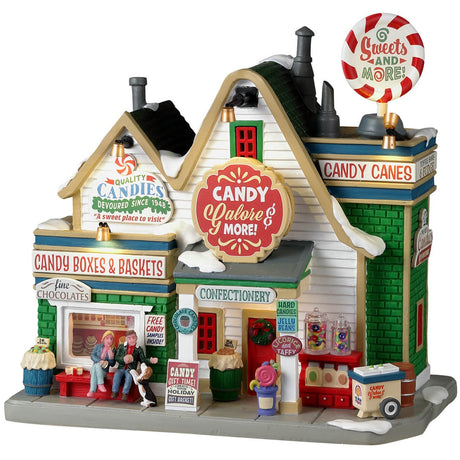 Resin lighted building for a Lemax display. Designed to look like a candy shop. There's a big peppermint sucker on top of the roof that reads "Sweets and More!" Other signs read "Quality Candies devoured since 1948. A sweet place to visit." There's a couple sitting on a red bench in front with a dog. There's also snow throughout