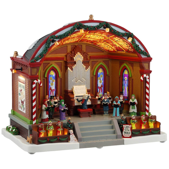 Resin lighted building for a Lemax display. Designed to look like a choir performing at a pavilion. There's a large organ, kids dressed like elves, two dogs, two peppermint candy cane accents, a decorated garland on the roof and a sign that reads "Joy to the World." The ceiling of the pavilion is decorated with different colored poinsettias.