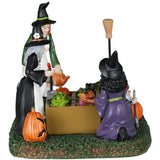 Resin table piece for Lemax display. Designed to look like three witches tending to a garden. There are pumpkins and radishes and lettuce and mushrooms and a black cat surveying the plants. There's a gnome statue holding a shovel.