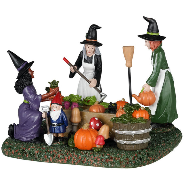 Resin table piece for Lemax display. Designed to look like three witches tending to a garden. There are pumpkins and radishes and lettuce and mushrooms and a black cat surveying the plants. There's a gnome statue holding a shovel.