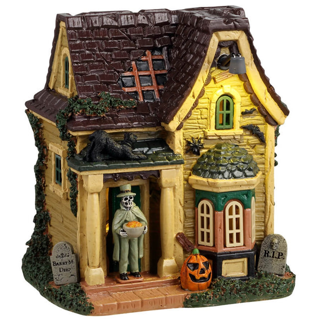 Resin building for a Lemax display. Designed to look like a run down house with moss growing on the side. Part of the roof is missing. There's a black cat on the roof and spiders. There's a skeleton wearing a lime green suit and cape handing out candy. There are tombstones in front of the house and a carved jack-o-lantern.